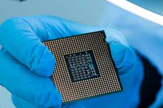 Gloved hand holding a computer processor chip with a grid of gold contacts on the underside.